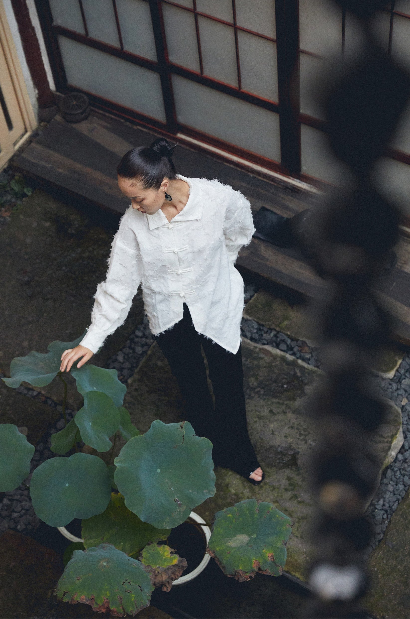 Woman in a white textured shirt and black pants standing next to a large green leafy plant indoors.