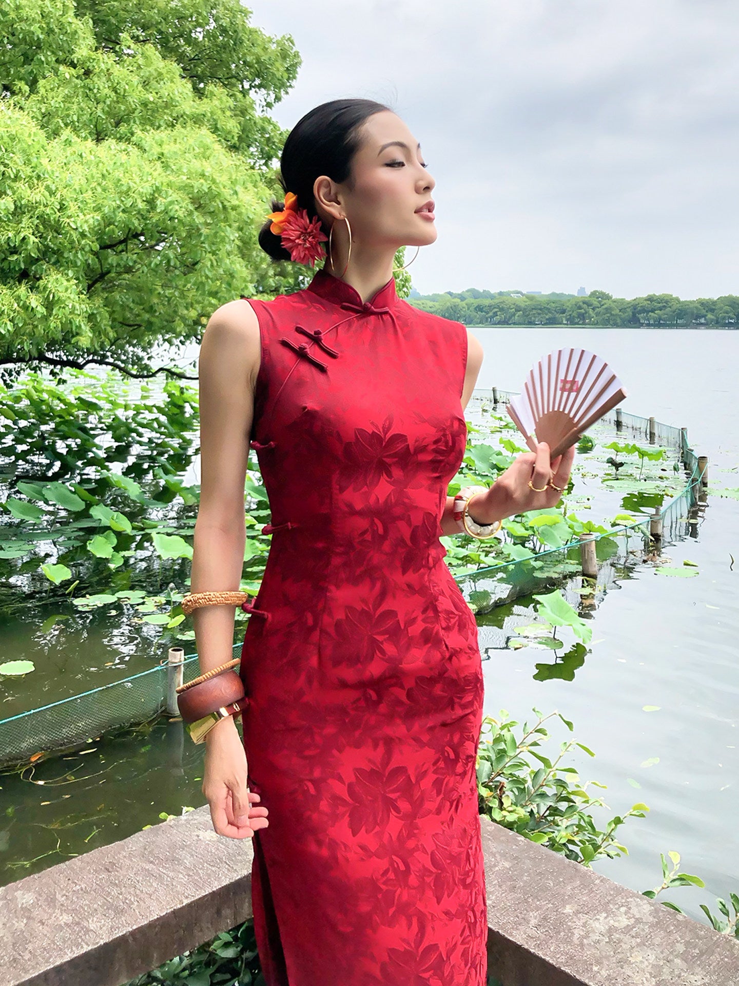Woman in a red dress holding a fan by a lake with greenery