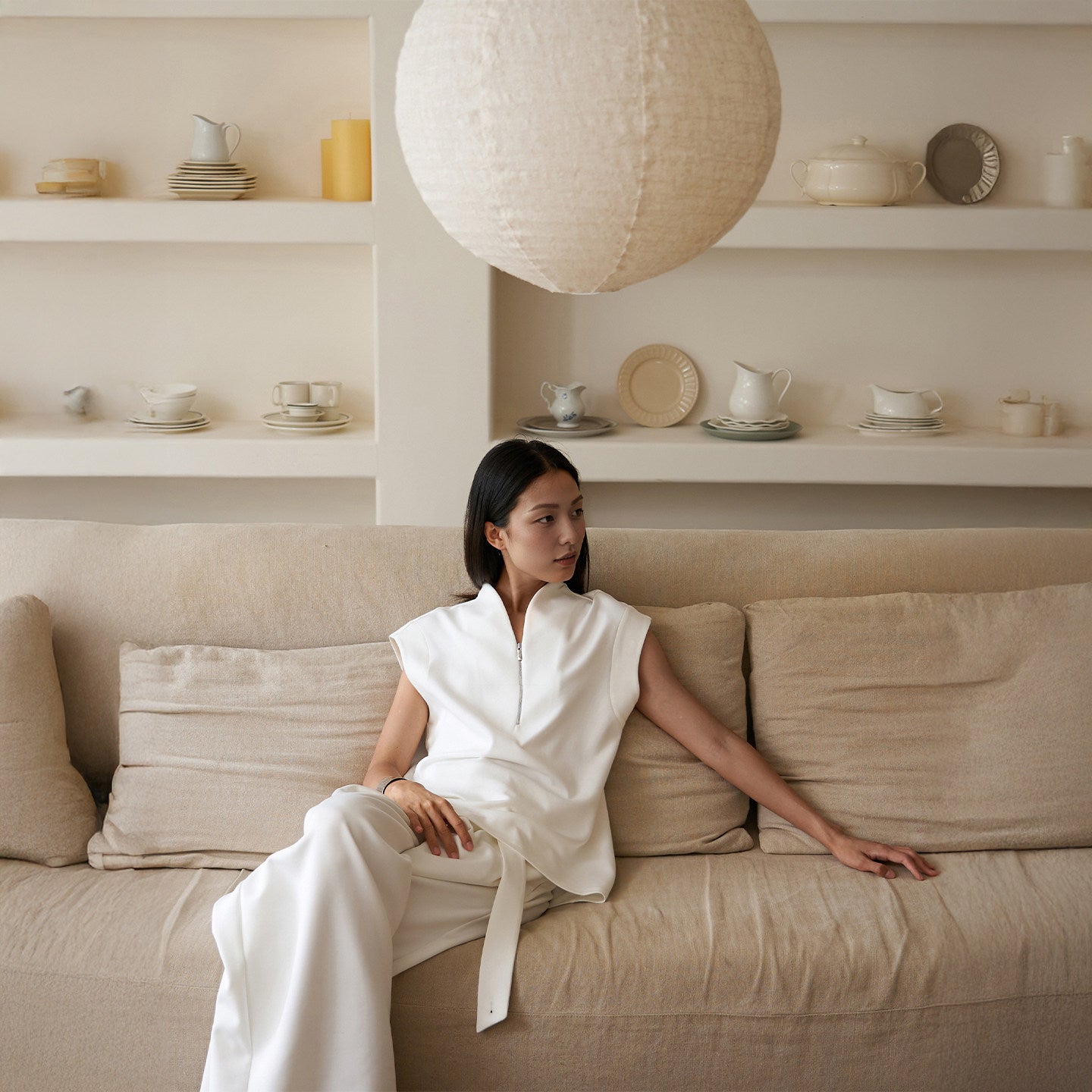 Woman in a white outfit sitting on a beige sofa in a room with shelves displaying ceramic items.