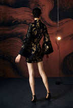 A woman's back standing against a textured wall, wearing a short, jacquard cheongsam dress with a stand collar neckline and full sleeves.