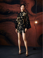 A woman standing against a textured wall, wearing a short, jacquard with slit cheongsam dress with a stand collar neckline and full sleeves.