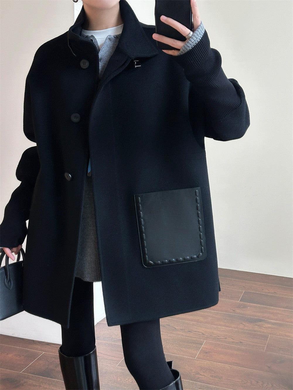 A person wearing a long black coat with a large front patch pocket, photographed indoors against a light wall and wood floor.