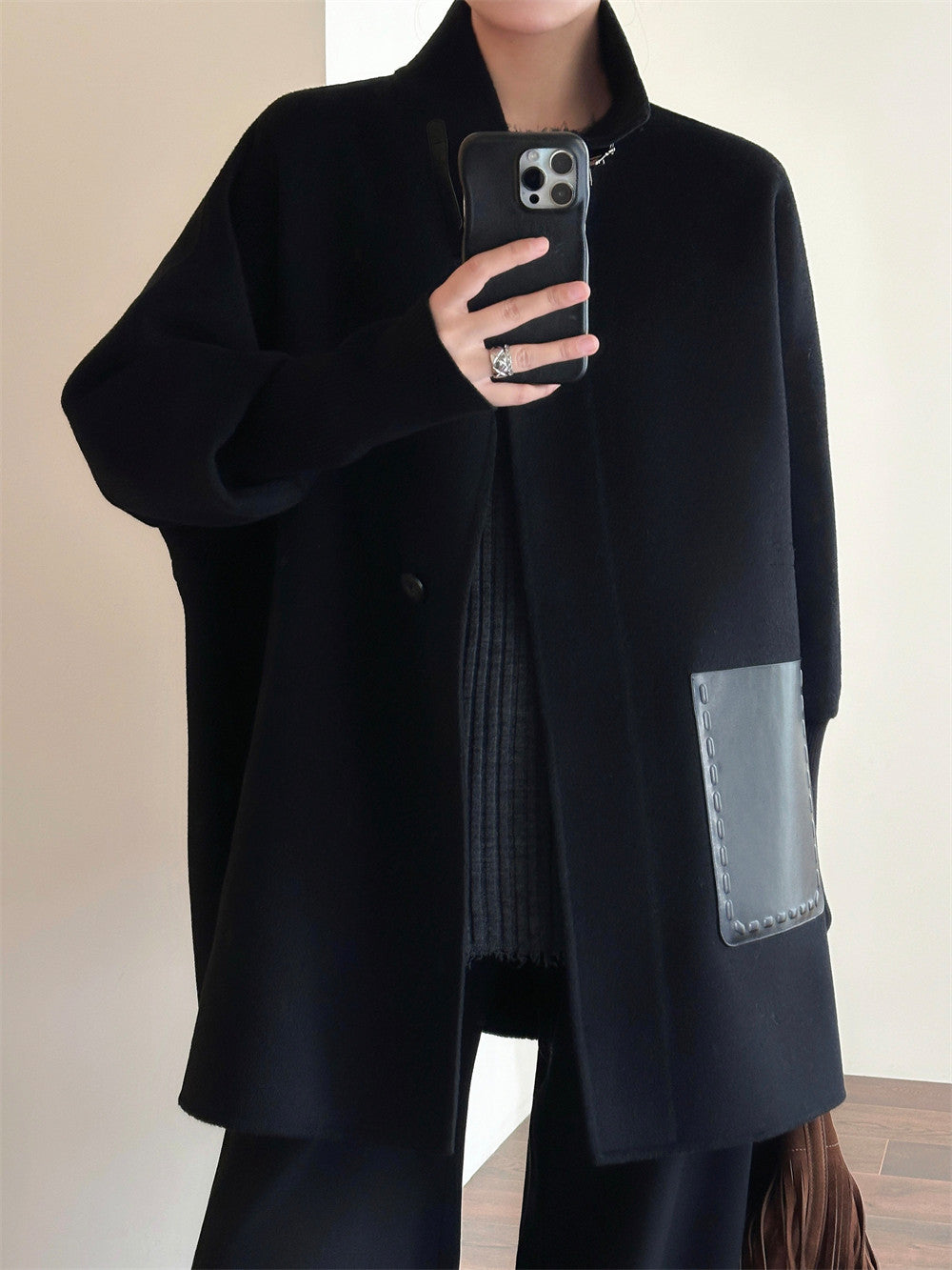 A person wearing a black oversized coat with a large patch pocket, taking a mirror selfie. The coat has a structured collar, long sleeves, and a contrasting dark gray pocket on the lower right side.
