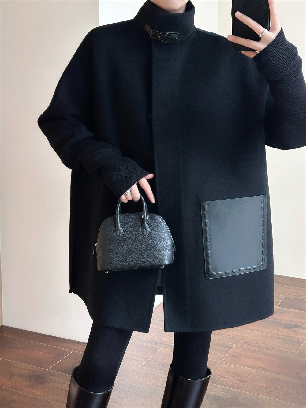 A person wearing an oversized black coat with a mock neck, black tights, black boots, and holding a structured black handbag in an indoor setting with a light wall and wood floor.