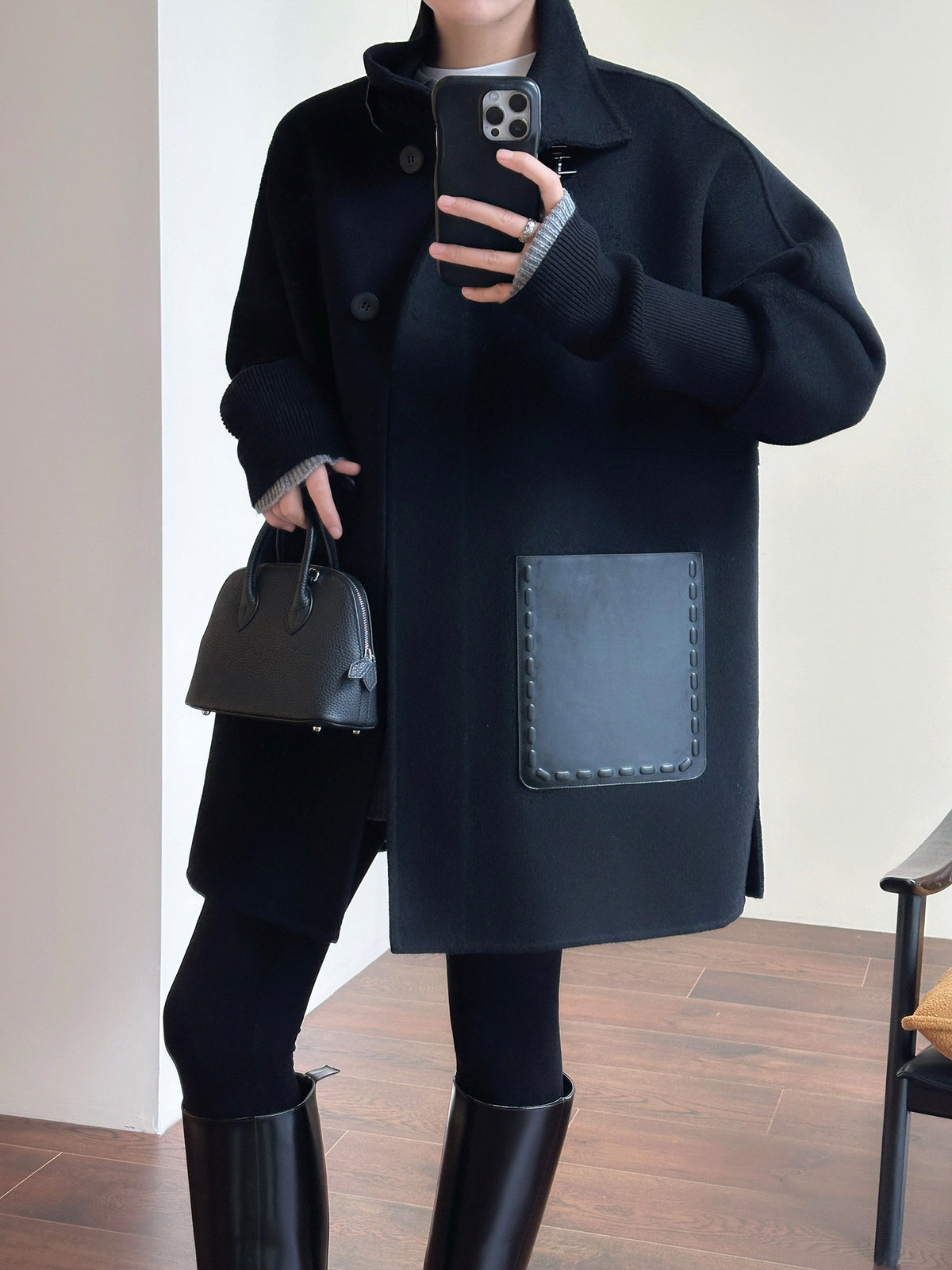 A person wearing an oversized black wool-blend coat with a large front patch pocket, paired with black leggings and tall black boots, holding a black handbag in an indoor setting.