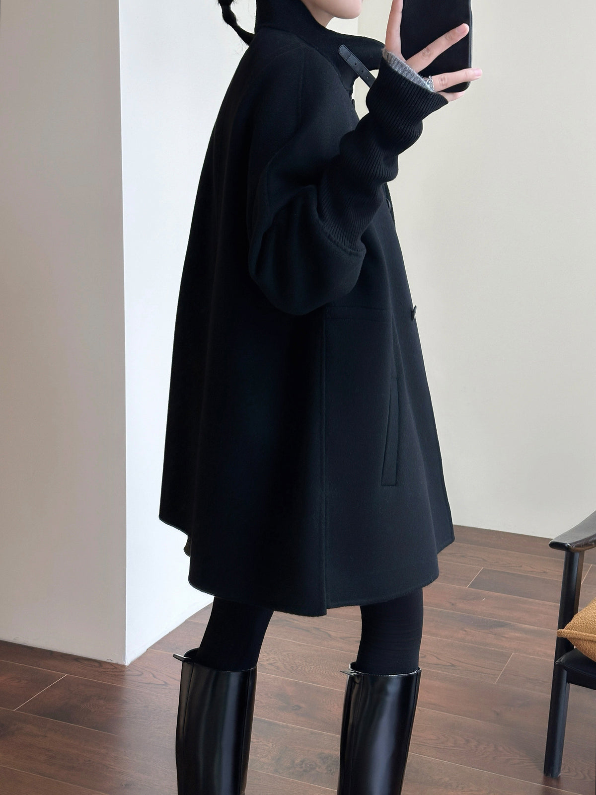 A person wearing a black oversized long-sleeve coat over a black turtleneck sweater, black leggings, and black knee-high boots, standing indoors against a light wall.