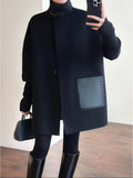 A person takes a mirror selfie wearing a black long coat with a high collar, a large patch pocket, and a single front button. The outfit includes a black turtleneck, black leggings, black knee-high boots, and a small black handbag, set against an indoor background with a light wall and wood floor.