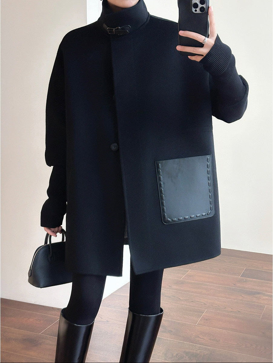 A person takes a mirror selfie wearing a black long coat with a high collar, a large patch pocket, and a single front button. The outfit includes a black turtleneck, black leggings, black knee-high boots, and a small black handbag, set against an indoor background with a light wall and wood floor.