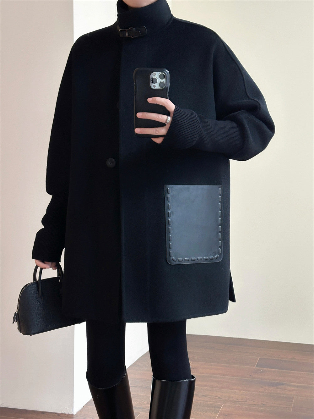 A person wearing an oversized black coat with a large rectangular patch pocket, black turtleneck, black leggings, and black knee-high boots, holding a black handbag and taking a mirror selfie indoors.
