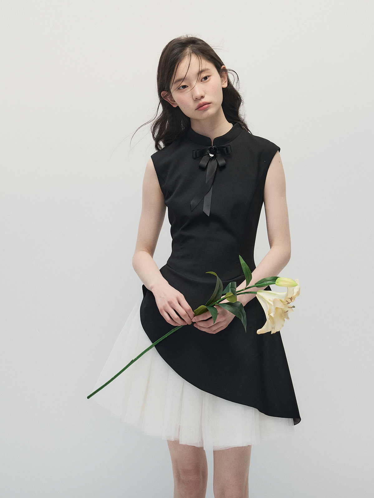 Woman wearing a black sleeveless top with a bow and white skirt holding flowers against a plain background