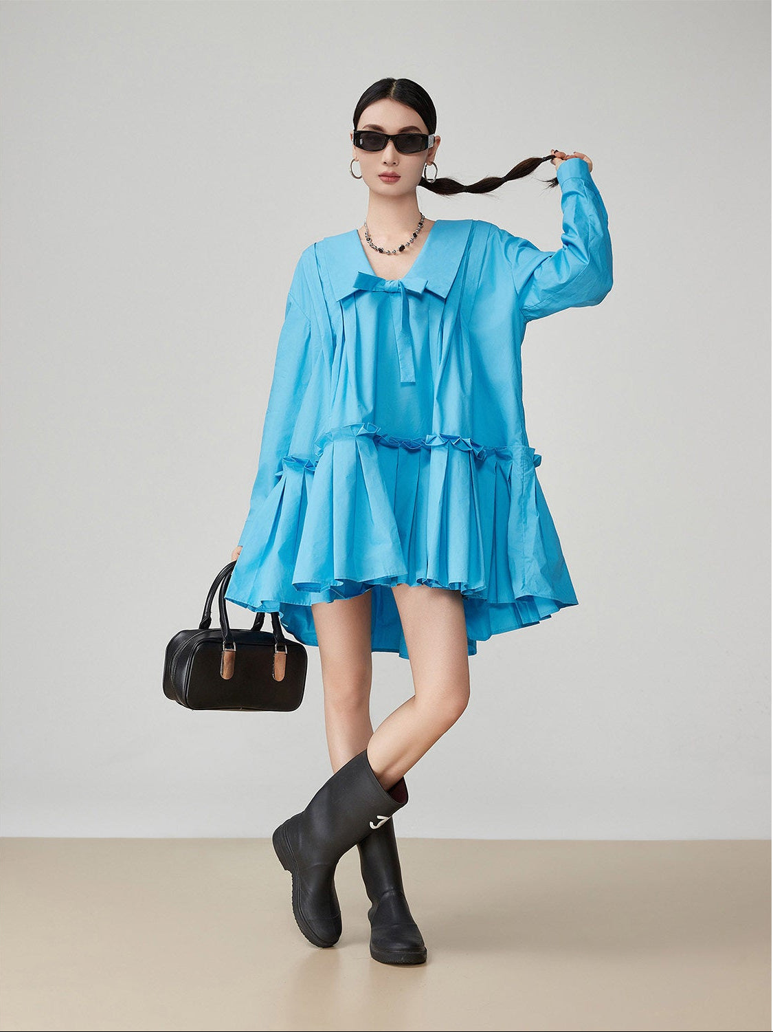 Woman wearing a blue dress with ruffles, black boots, and holding a black handbag against a plain background.