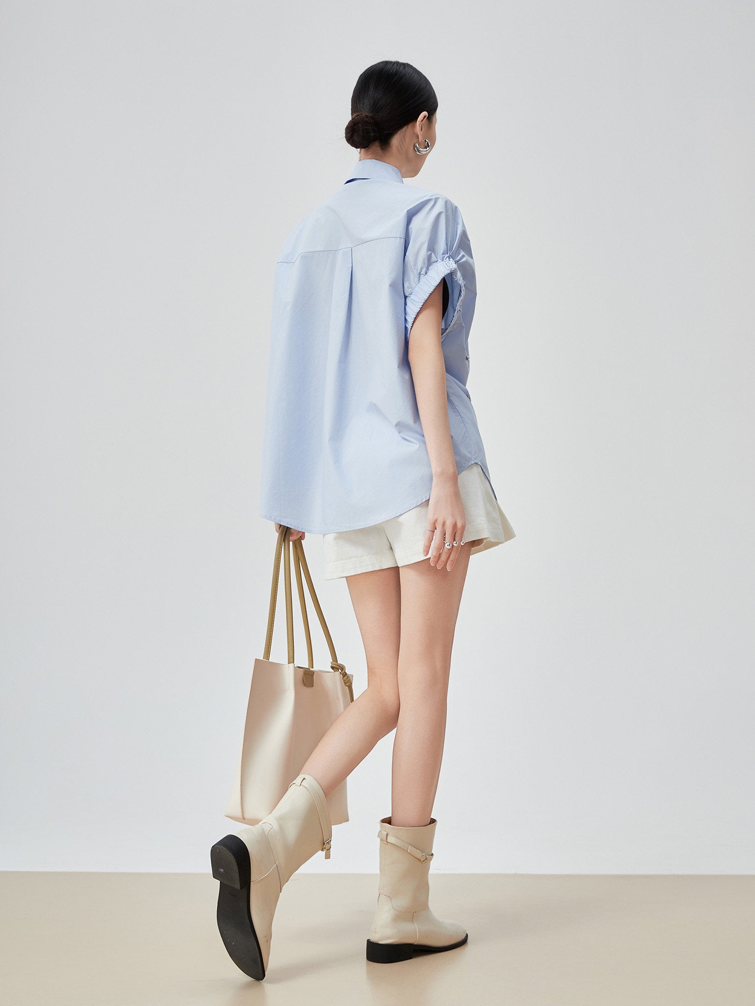 Woman wearing a light blue shirt, white shorts, and beige boots, holding a cream-colored tote bag.