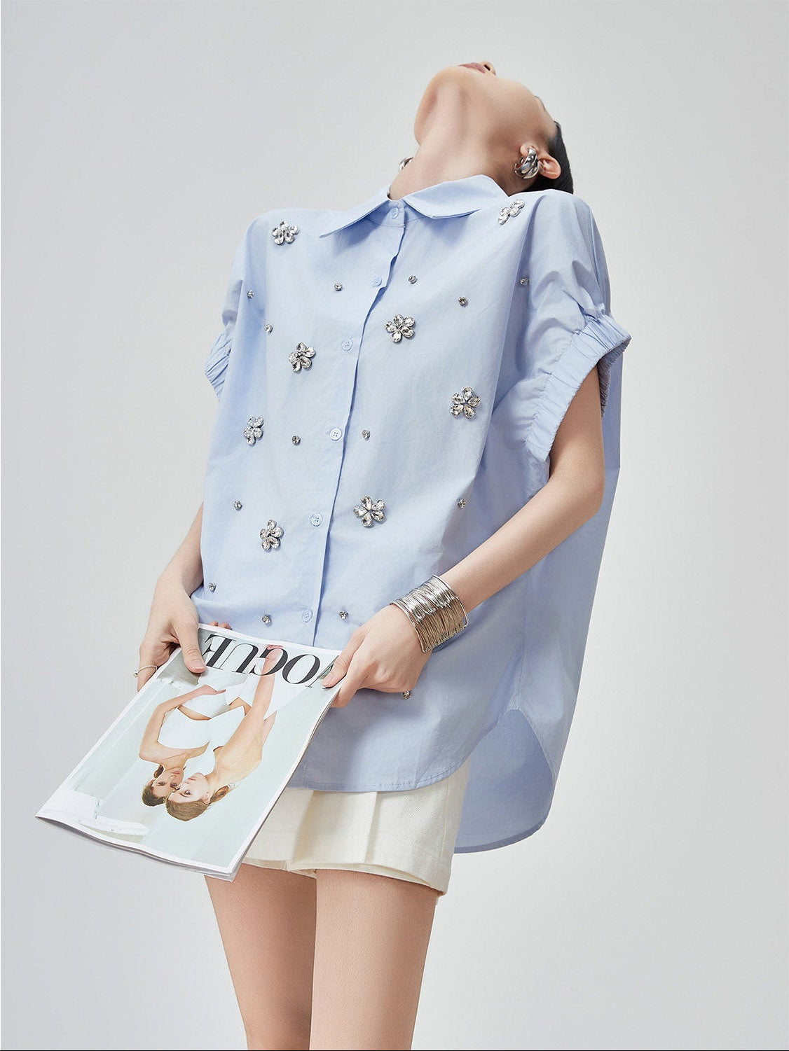 Woman wearing a light blue shirt with floral embellishments holding a magazine on a gray background