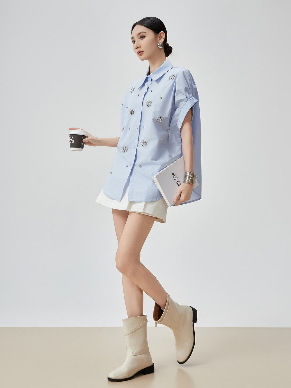 Woman wearing a light blue shirt with floral patterns, white skirt, and beige ankle boots on a plain background.