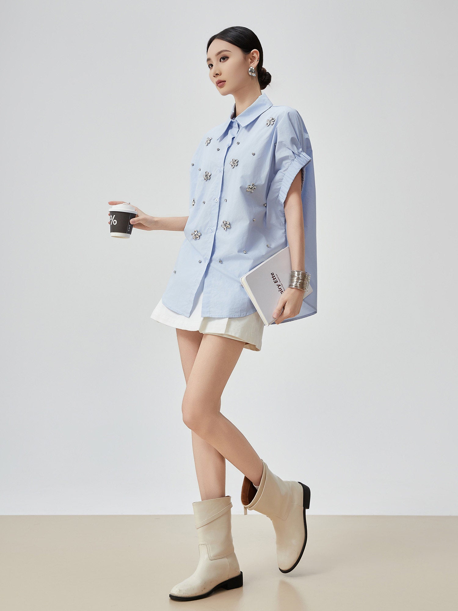 Woman wearing a light blue shirt with floral patterns, white skirt, and beige ankle boots on a plain background.