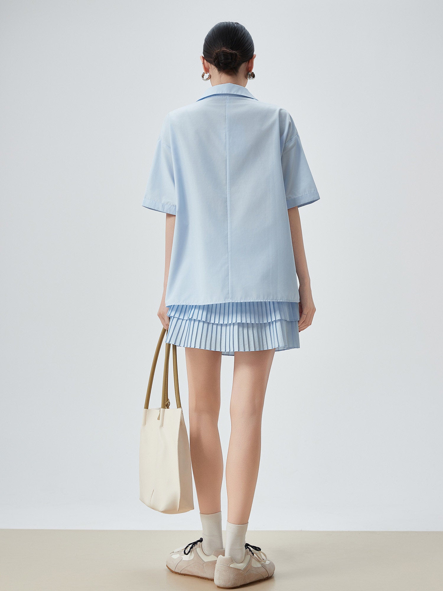 Person wearing a light blue outfit with a pleated skirt, holding a beige tote bag on a plain background