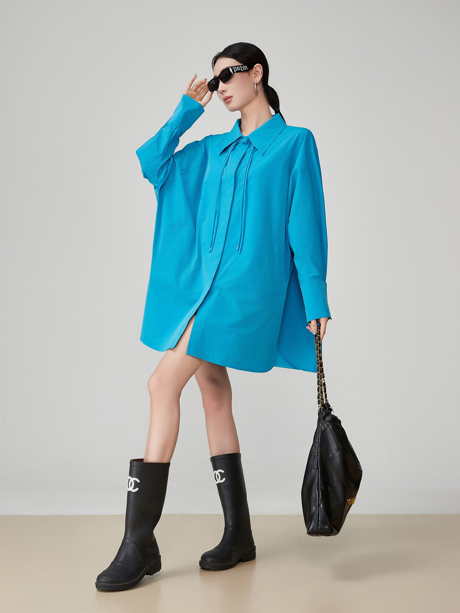 Woman wearing a blue oversized shirt, black rain boots, and holding a black bag against a plain background