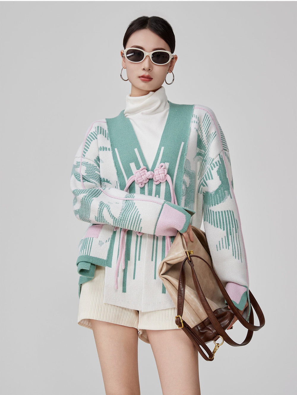 Woman wearing a patterned cardigan with a white turtleneck and beige shorts, holding a brown handbag.