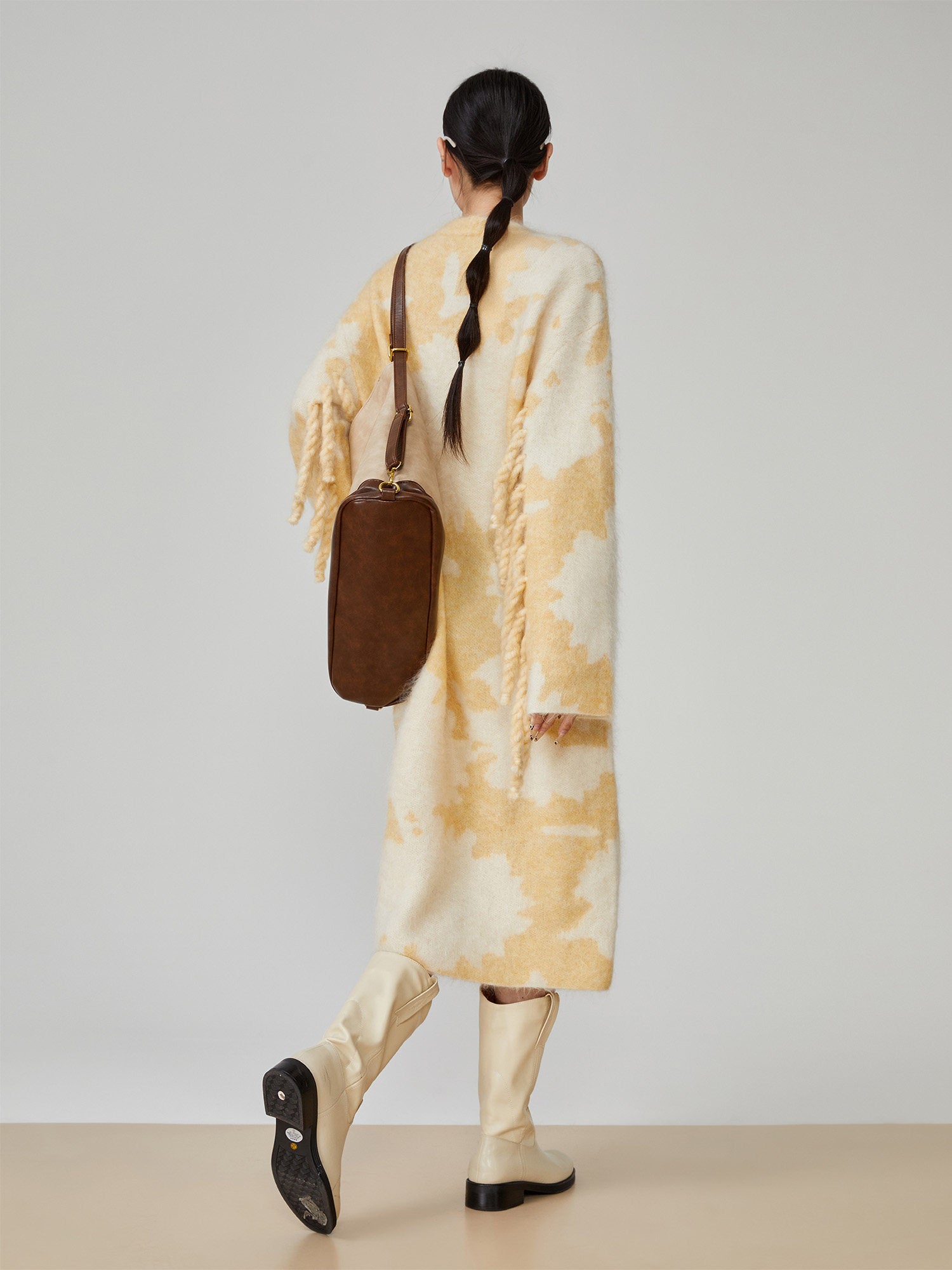 Person wearing a long, light-colored coat with a brown bag on a plain background