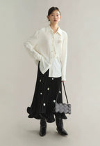 Woman wearing a white blouse, black pleated skirt, and black boots with a gray bag on a light gray background