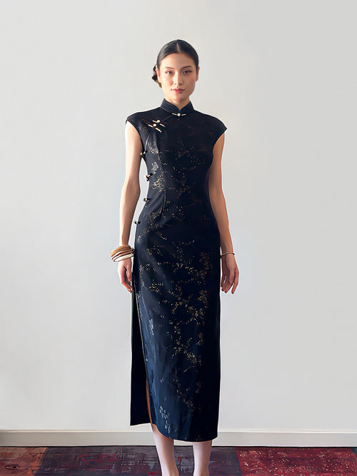 Woman wearing a black dress with gold patterns against a white wall.