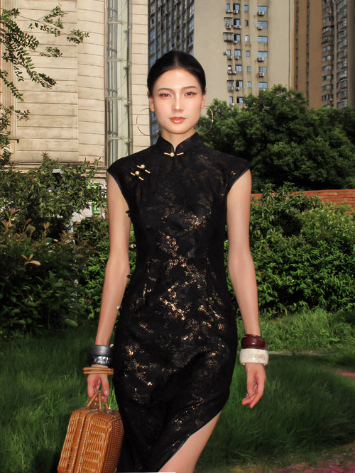 Woman in a black dress with gold patterns walking outdoors with greenery and buildings in the background