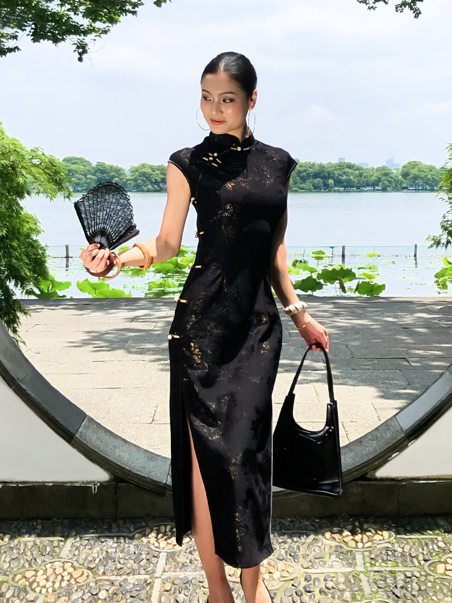 Woman in a black dress holding a fan and bag by a waterfront
