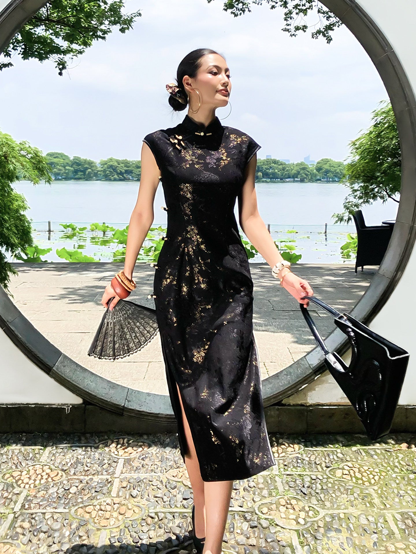 Woman in a black lace dress standing outdoors near a circular structure with water and trees in the background.