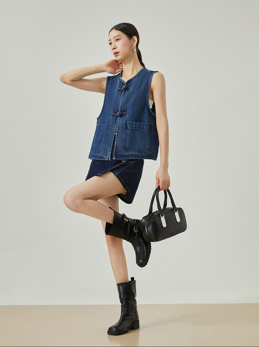 Woman wearing a denim vest and skirt with black boots and handbag on a plain background