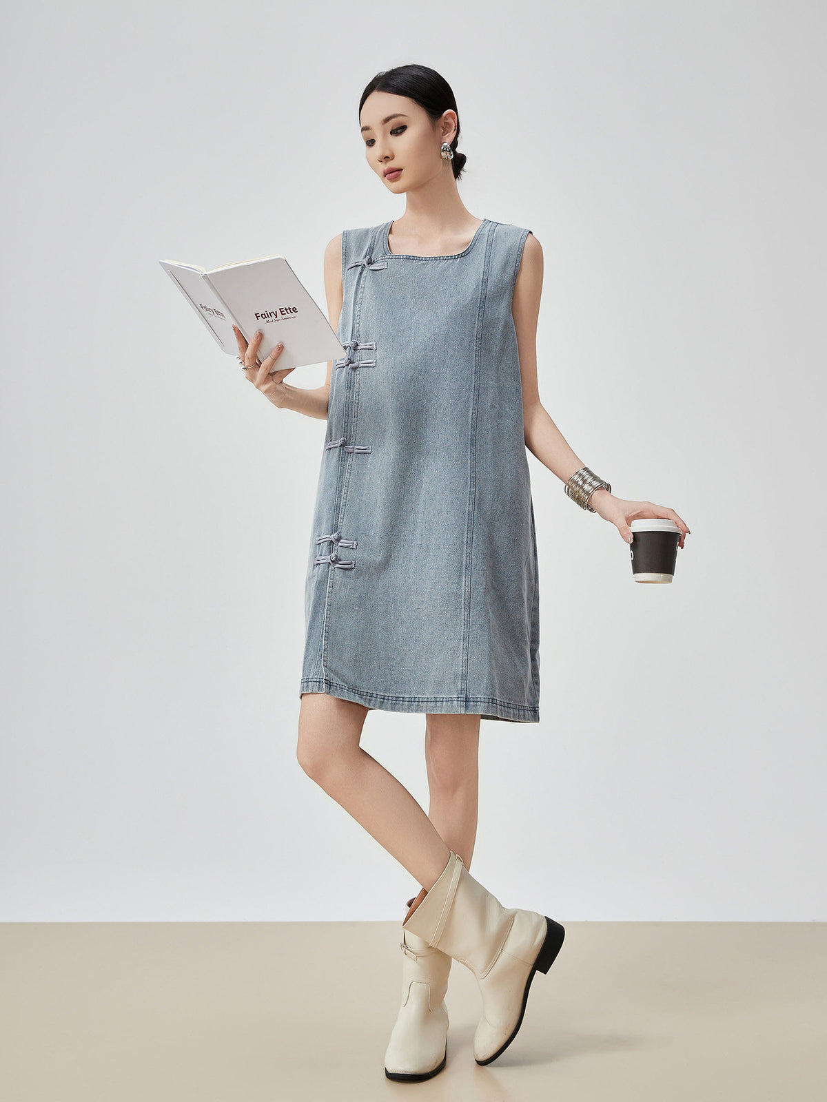 Woman wearing a denim dress holding a coffee cup and a book on a plain background
