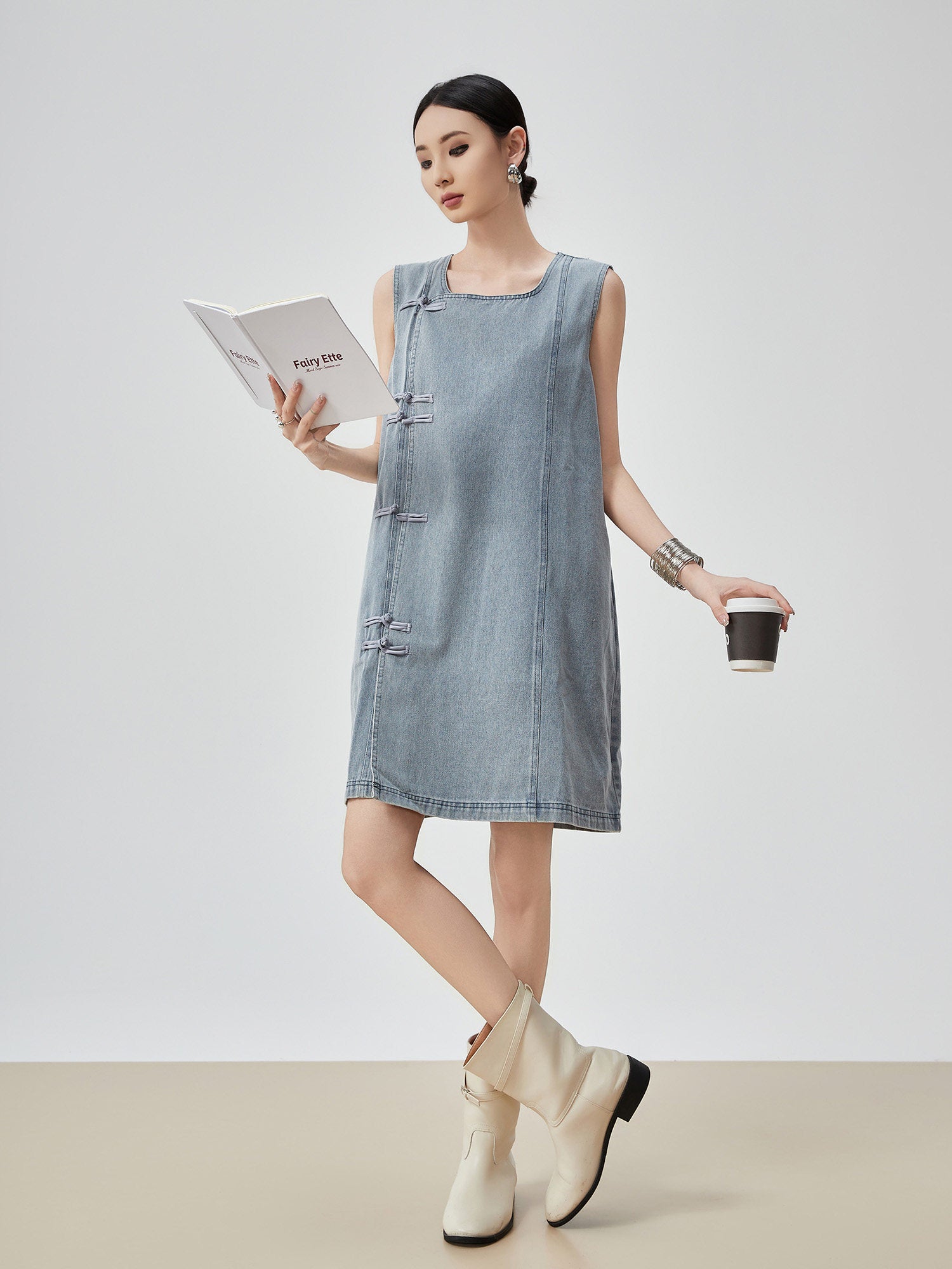 Woman wearing a denim dress holding a coffee cup and a book on a plain background