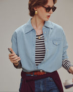 Woman wearing a light blue denim shirt over a striped top, holding a phone, with a neutral background.