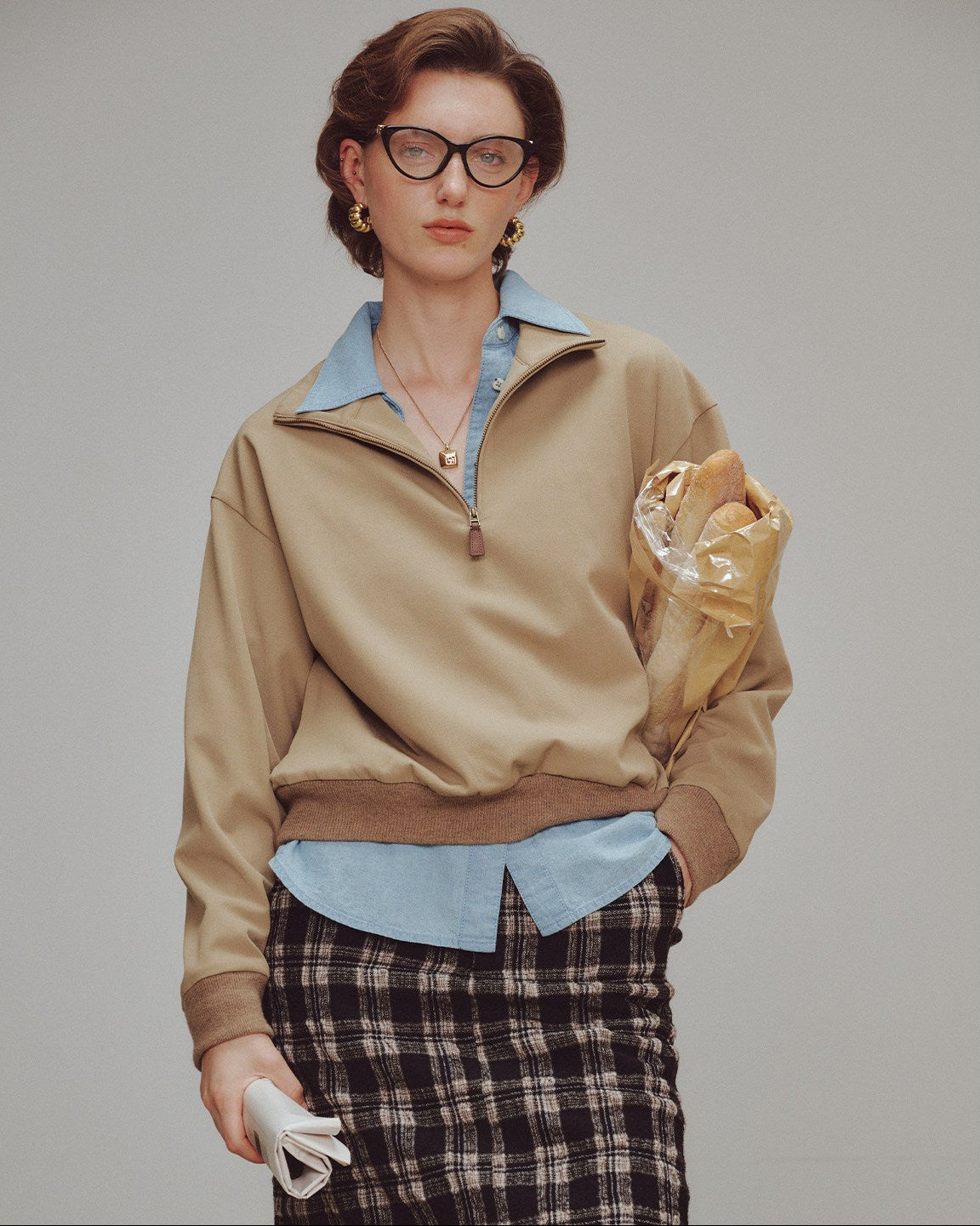 Woman wearing a beige jacket over a blue shirt with plaid pants, holding a gold bag.