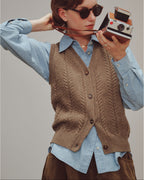 Woman wearing a brown knitted vest over a light blue shirt, holding a vintage camera.