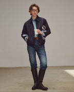 Woman wearing a dark jacket, light shirt, jeans, and black boots holding a white cup.