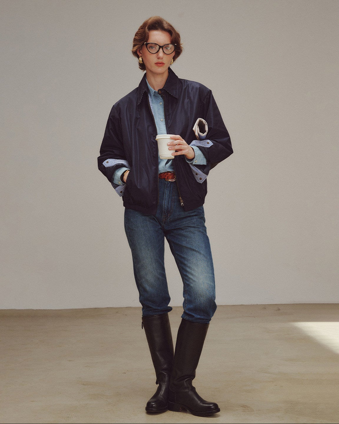 Woman wearing a dark jacket, light shirt, jeans, and black boots holding a white cup.