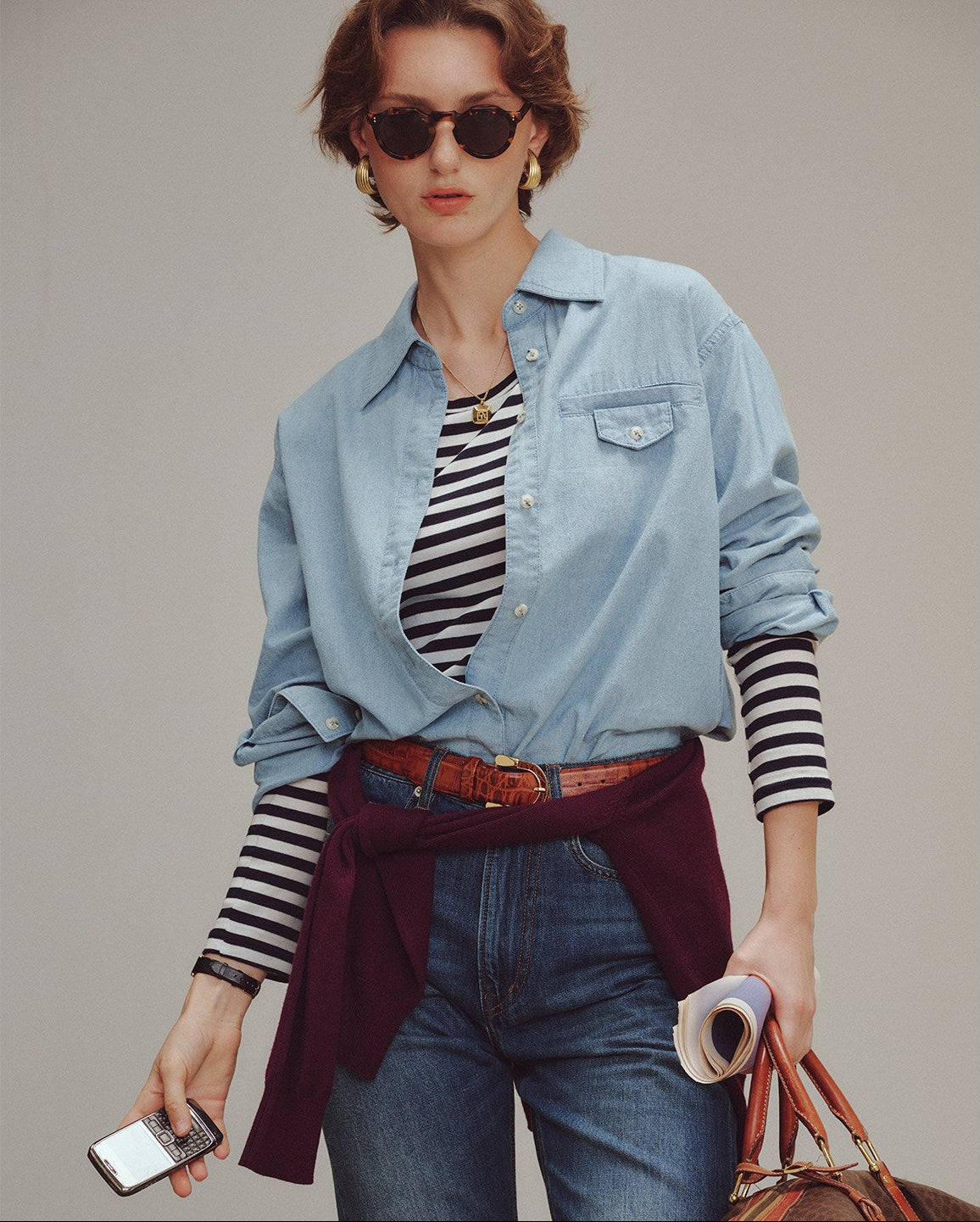 Woman wearing a denim shirt over a striped top with jeans, holding a phone and a bag.