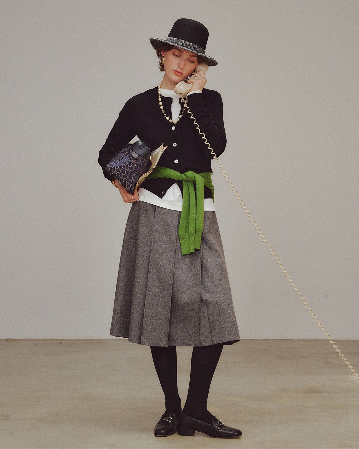 Woman in vintage outfit with hat, skirt, and coat holding a phone receiver against a plain background