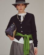 Woman wearing a black cardigan, white shirt, grey skirt, and green belt, holding a  handbag.