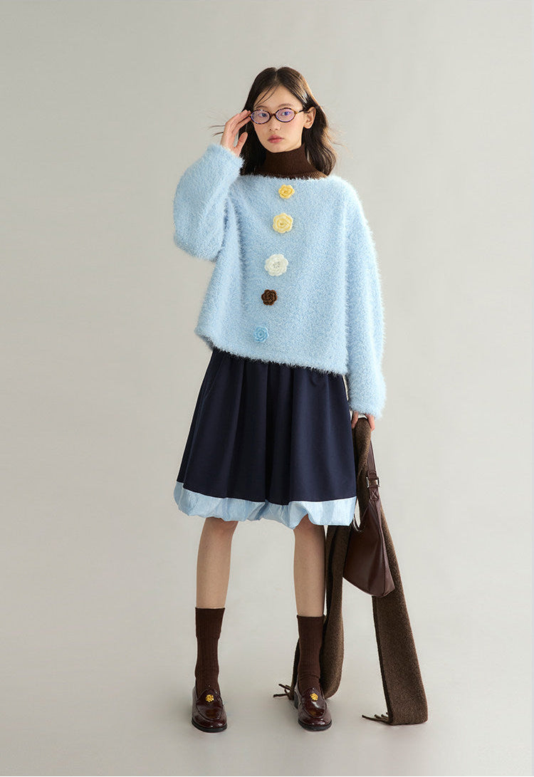 A model stands against a light gray studio background wearing a fuzzy light blue cropped sweater with decorative flower-like buttons, a dark navy skirt with a light blue hem, brown socks, and brown shoes, while holding a large brown bag.