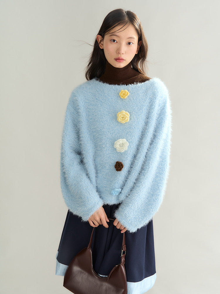 A model wearing a light blue fuzzy knit sweater with large 3D fabric flowers down the front, styled over a dark brown mock neck top and navy blue skirt, holding a brown leather handbag against a plain light gray background.
