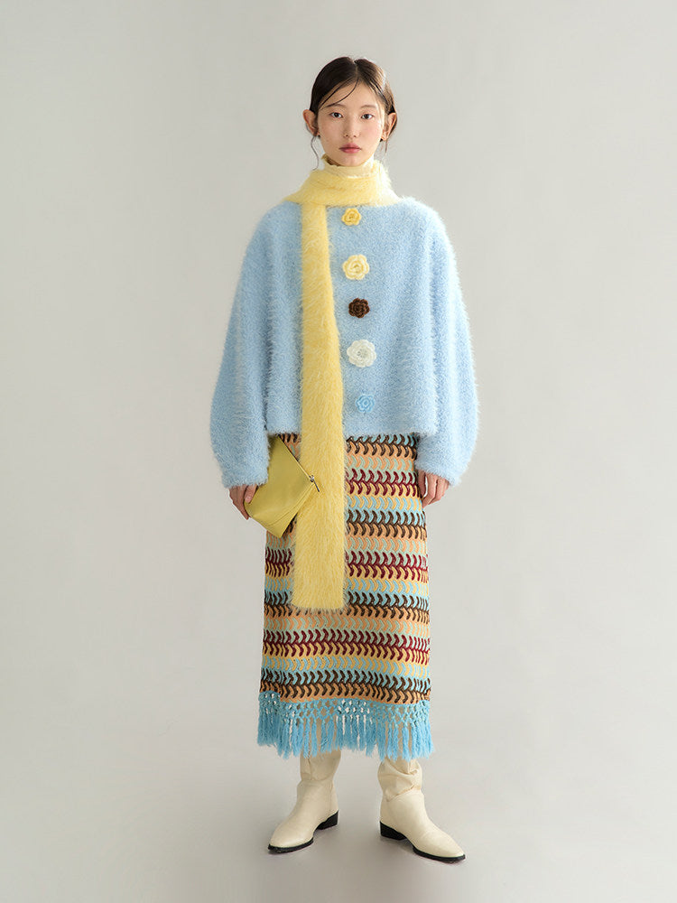 A model stands against a light gray studio background wearing a light blue fuzzy cropped cardigan with yellow buttons, a bright yellow scarf and clutch, and a multicolor patterned knit skirt with blue fringe, paired with cream ankle boots.
