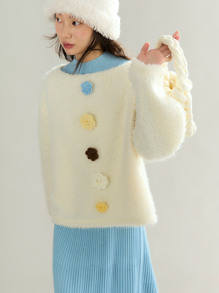 A model wearing a fuzzy cream-colored cardigan sweater with decorative flower buttons in blue, yellow, white, and brown, styled with a light blue ribbed top and skirt, a white fluffy hat, and holding a matching cream bag.