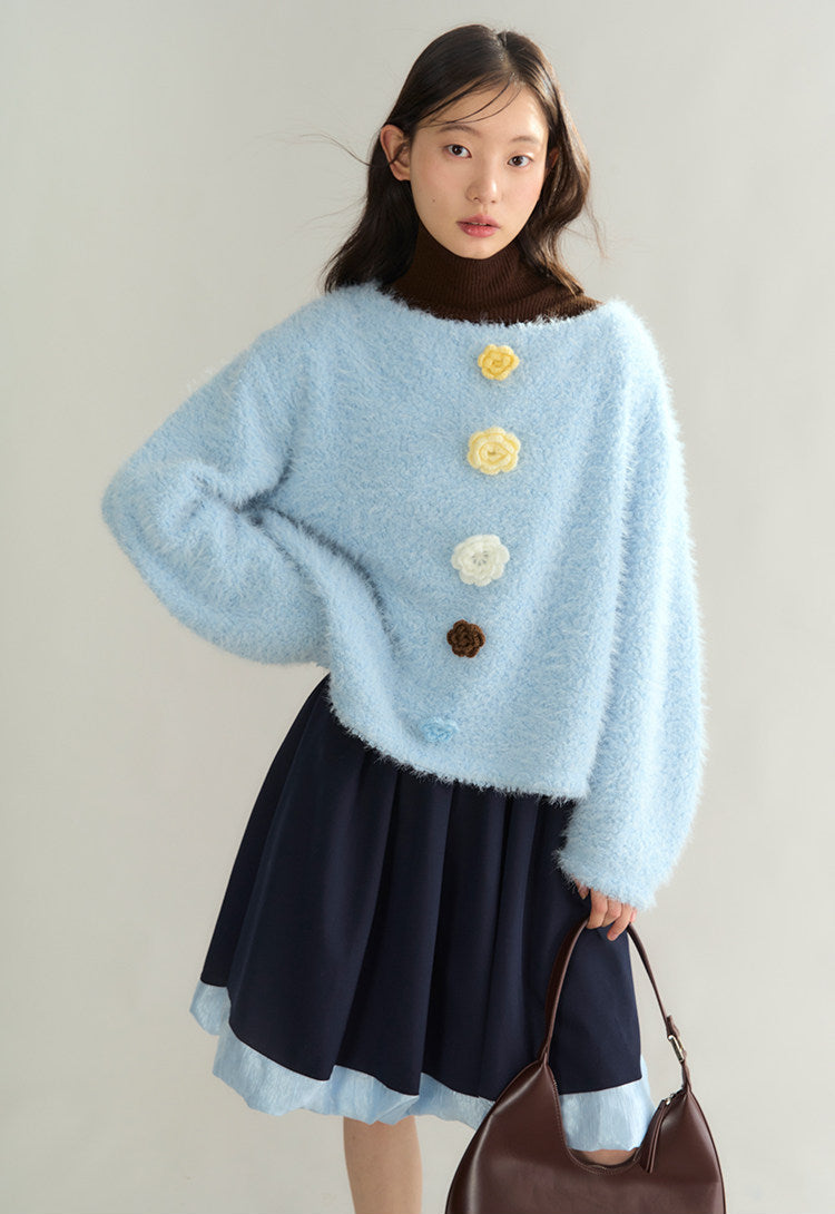 A model wearing a light blue fuzzy cropped sweater with 3D flower buttons, a dark navy pleated skirt, and holding a brown leather shoulder bag against a plain light gray background.