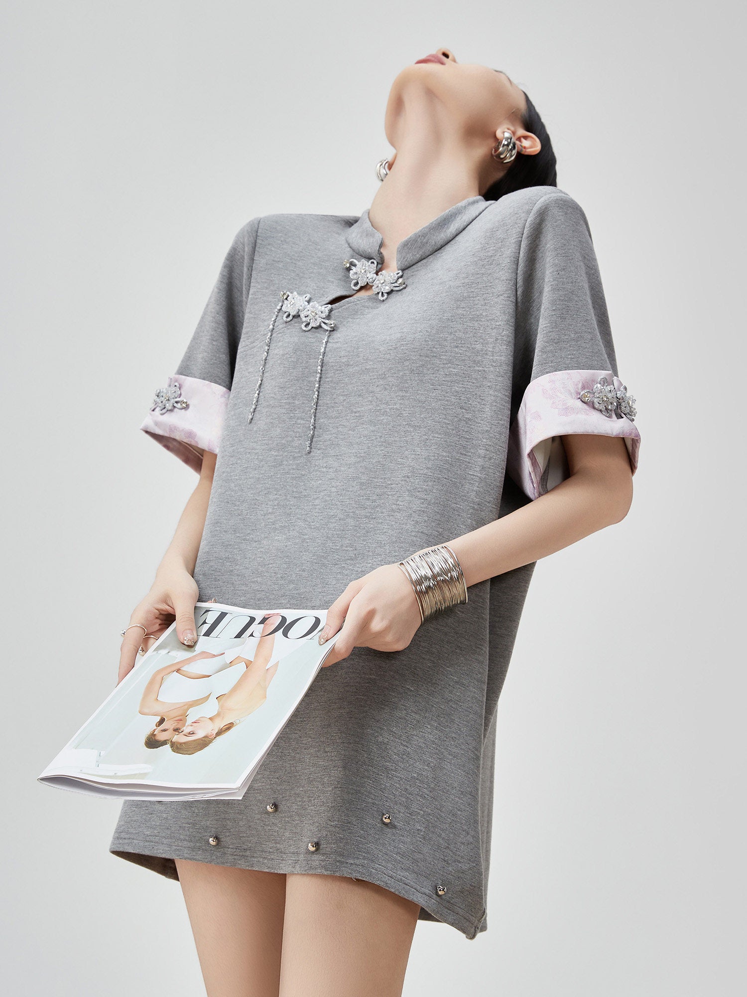 Woman wearing a gray dress with embellishments, holding a magazine on a light gray background