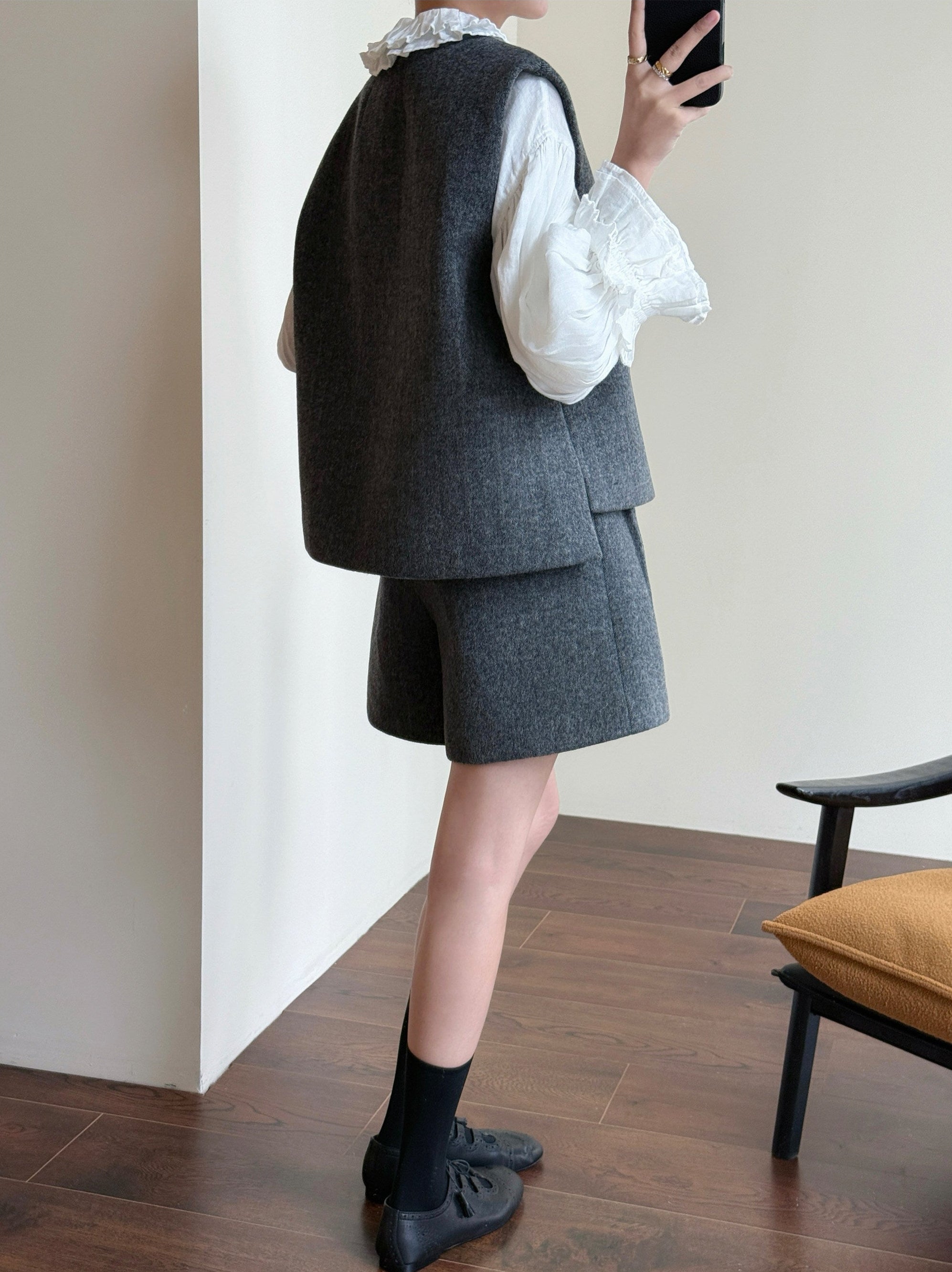 A person shown from the back wearing a dark gray sleeveless vest and matching shorts over a white blouse with ruffled sleeves, paired with black socks and black shoes, standing indoors near a chair.
