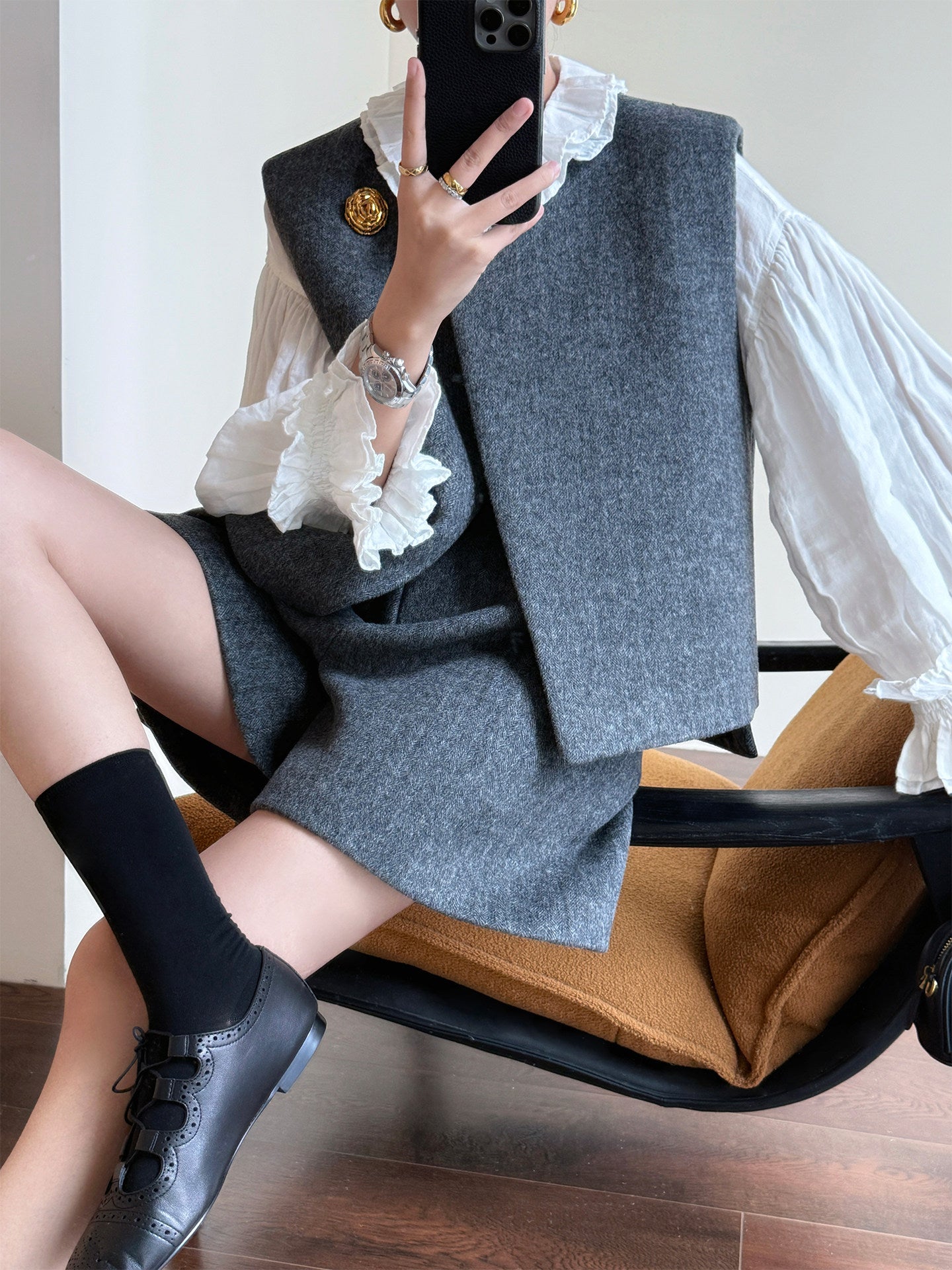 A person seated in a chair taking a mirror selfie while wearing a gray sleeveless tailored vest and matching gray skirt over a white ruffled blouse, with black socks and black lace-up shoes.