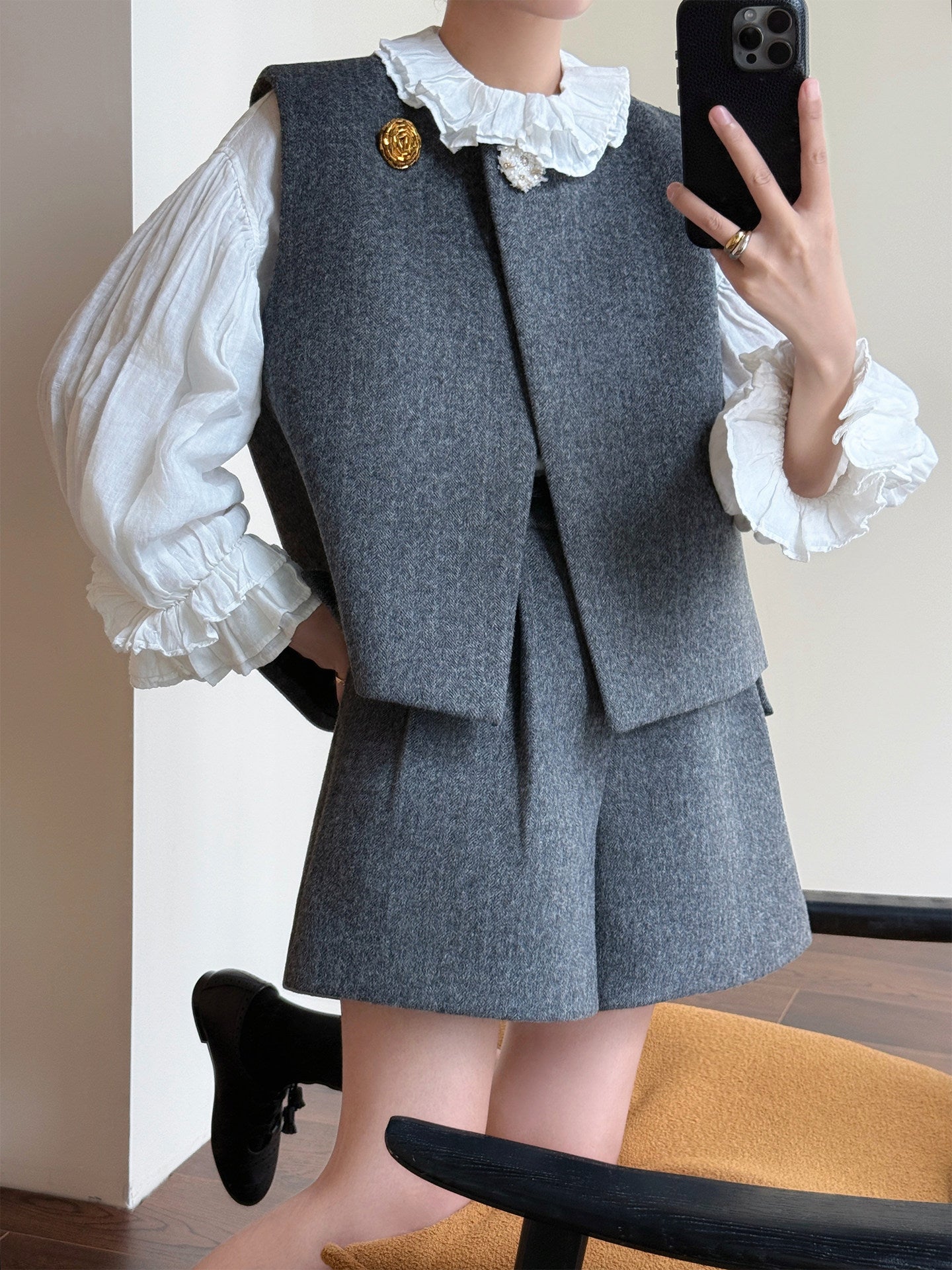 A person takes a mirror selfie wearing a gray tweed vest and matching shorts set with a white ruffled blouse. The outfit is styled with a gold brooch and a black shoe.