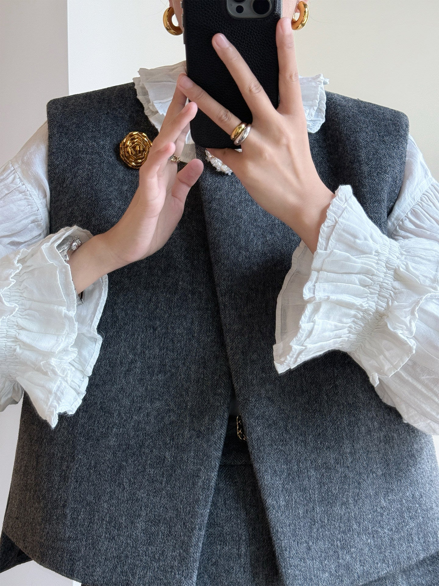 A person taking a mirror selfie wearing a dark gray wool vest over a white ruffled blouse, accessorized with gold rings and earrings.