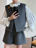 A person takes a mirror selfie showing a gray two-piece vest and shorts set with a white ruffled blouse. The vest has a gold decorative pin on the chest.